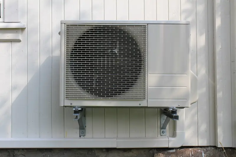 Outdoor heat pump unit hung high on steel wall brackets above the snow line on a pale board-and-batten house, its line set tucked in a cover along the foundation