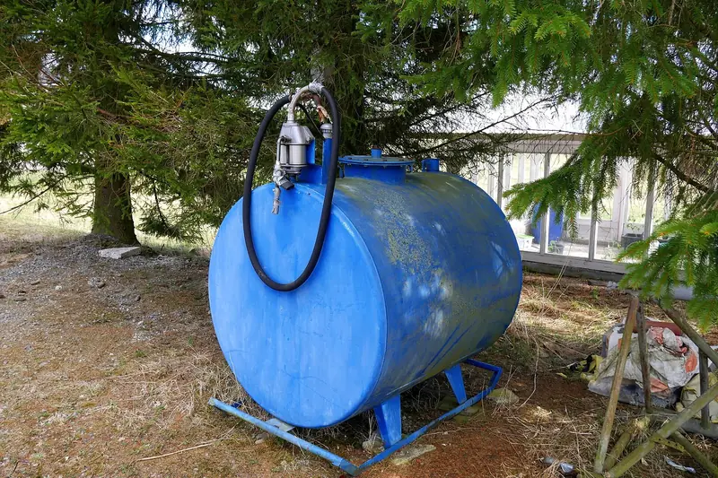 Blue horizontal steel fuel tank on a steel frame under spruce trees, with a hand pump and hose fixed to one end