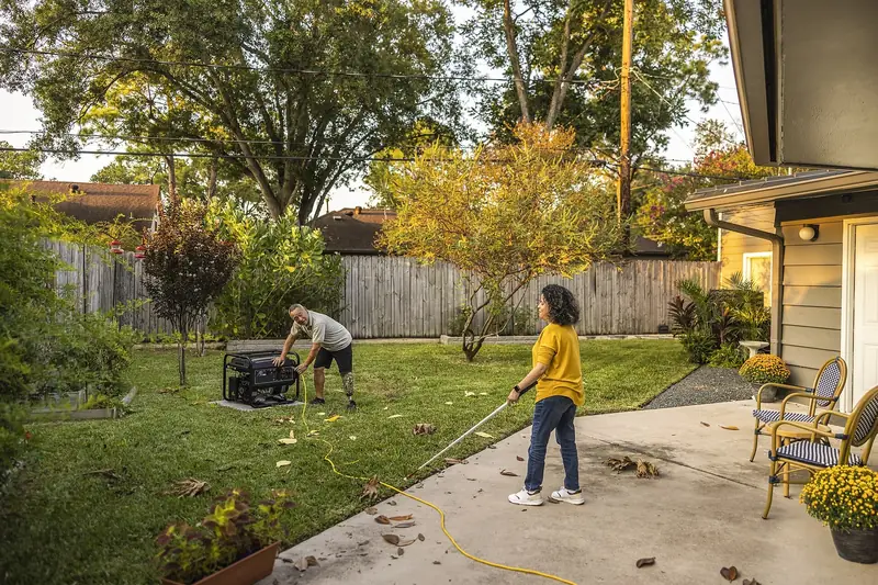 Man pulling the starter on a portable generator set on a concrete pad out in a fenced backyard, an extension cord laid across the grass toward the patio