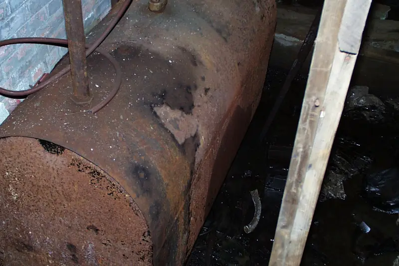 Close look at the end of an old steel oil tank crusted with orange rust and deep pits, lit dimly in a cellar